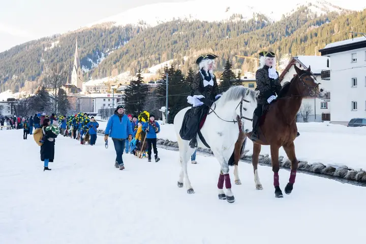Zwei Füerriiter reiten vor dem Umzug, dahinter Davos mit dem Kirchturm
