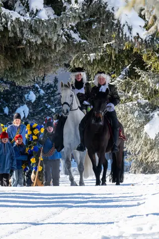 Die Füerriiter reiten unter Tannen, dahinter Kinder in blauen Jacken