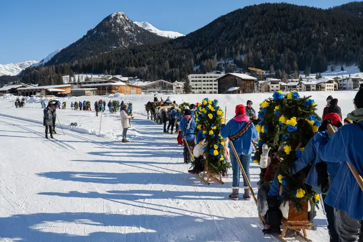 Eine lange Reihe von Rennbänna zieht über die Schneefläche, dahinter Berge