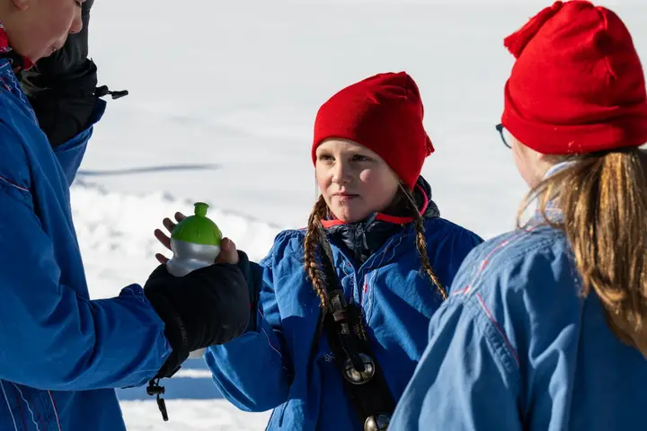 Kinder in blauen Jacken, eines mit roter Mütze, bekommen etwas zu trinken