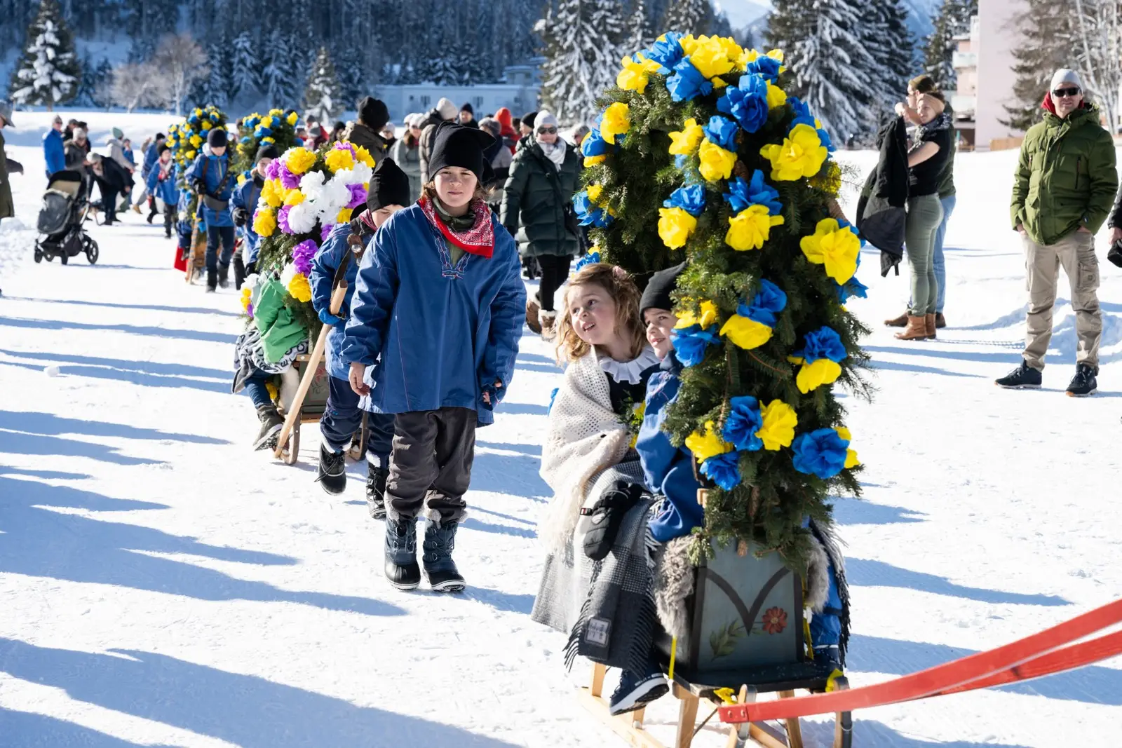 Kinder in blauen Jacken gehen in einer langen Reihe neben mit Papierrosen geschmückten Rennbänna über den Schnee, rechts ein hoher Rosenkranz