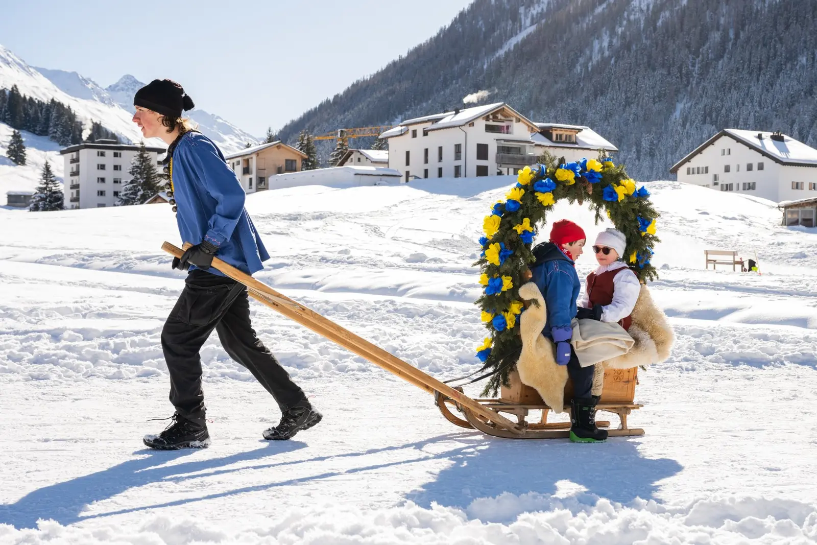 Ein Kind in blauer Jacke hält die langen Holme einer Rennbänna und zieht sie über den Schnee, zwei Kinder sitzen unter dem Rosenbogen