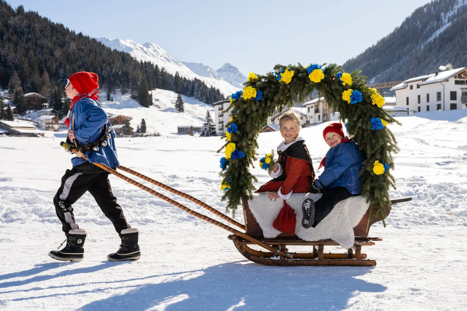 Ein Kind mit roter Mütze zieht die Rennbänna an Leinen durch den Schnee, zwei Kinder sitzen unter dem Rosenbogen
