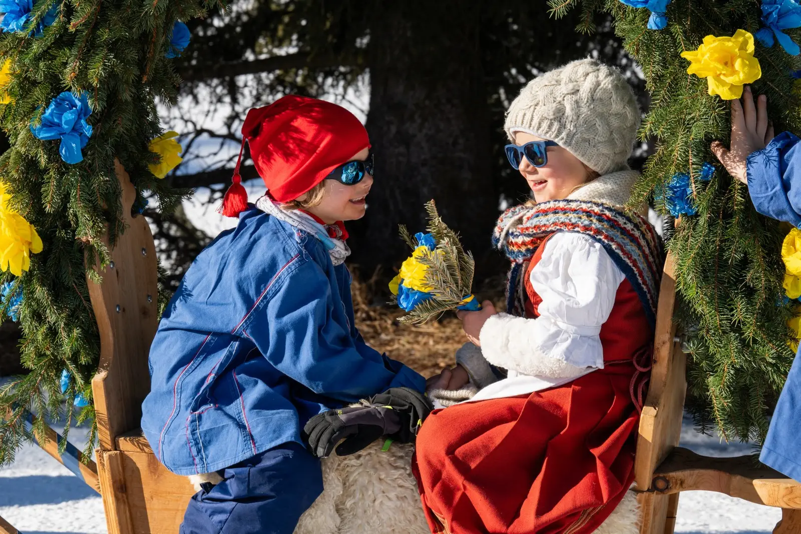 Ein Mädchen in Tracht mit rotem Rock sitzt im Schlitten zwischen Papierrosen und schaut ein Kind in blauer Jacke an