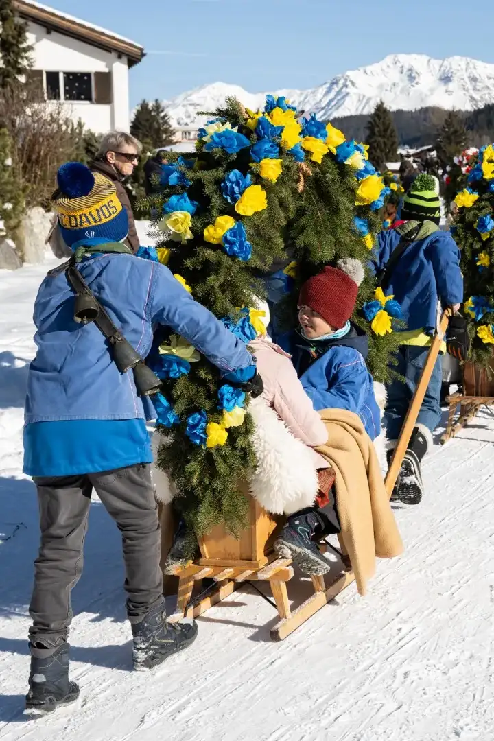 Kinder in blauen Jacken ziehen und schieben mit Papierrosen geschmückte Schlitten in einer Reihe über den verschneiten Weg, dahinter die Davoser Berge